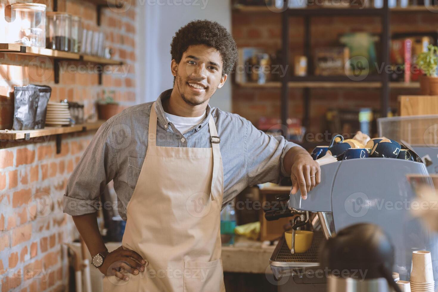 Friendly meeting in favorite cafe in morning. Smiling young african american barista guy in apron stands near espresso machine at bar and looks at camera in interior of cafeteria, empty space photo