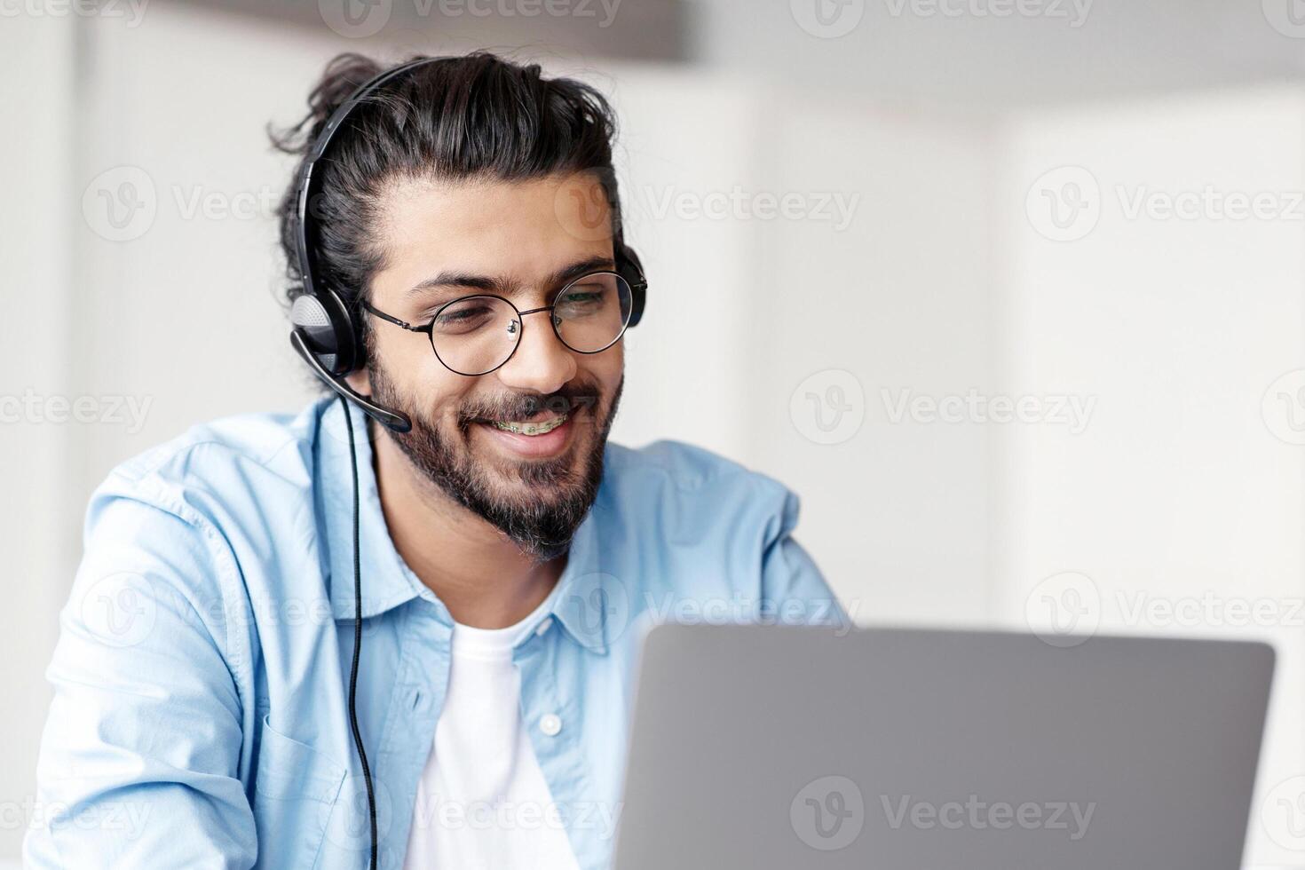 Hotline Operator. Portrait Of Indian Customer Support Manager Man In Headset Working With Laptop Computer In Office, Consulting Clients, Western Guy Sitting At Desk And Smiling, Free Space photo