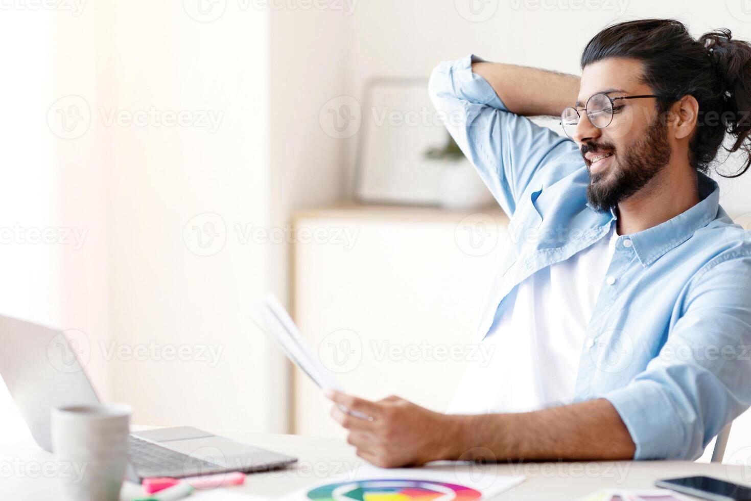 Young eastern designer man holding and looking at paper colour swatches, working at desk in home office, indian male entrepreneur choosing color samples for interior design project, free space photo