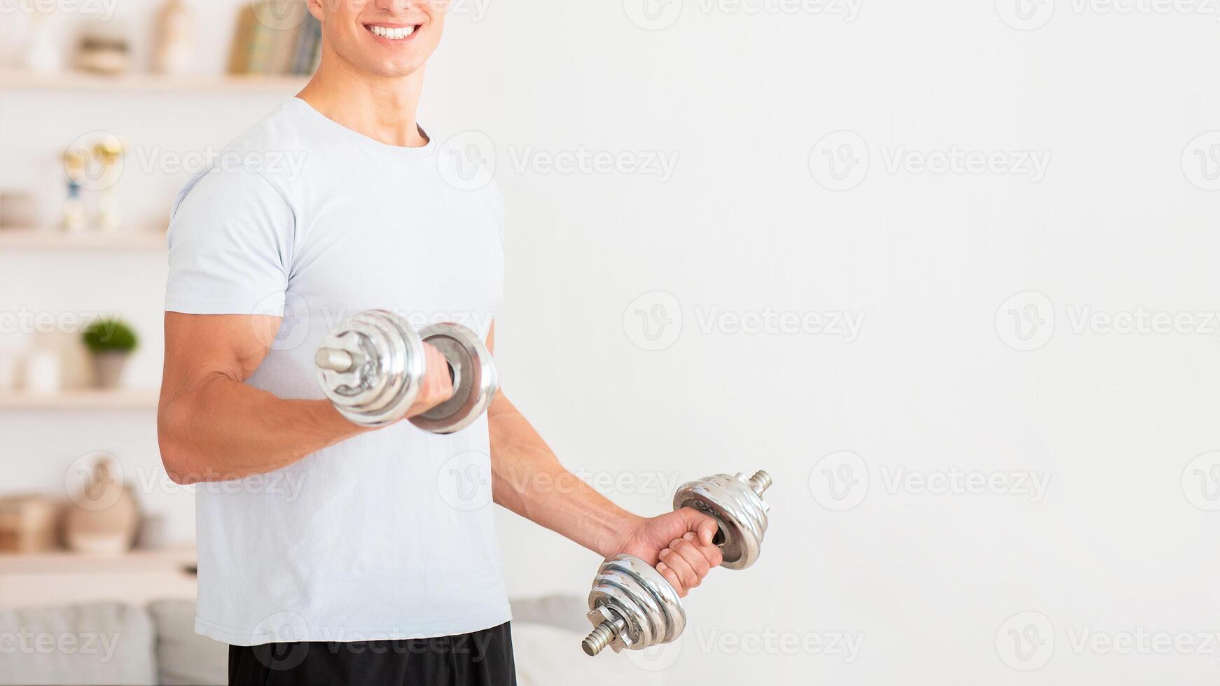 Strength training at home during quarantine. Smiling handsome guy lifts dumbbells in living room interior photo