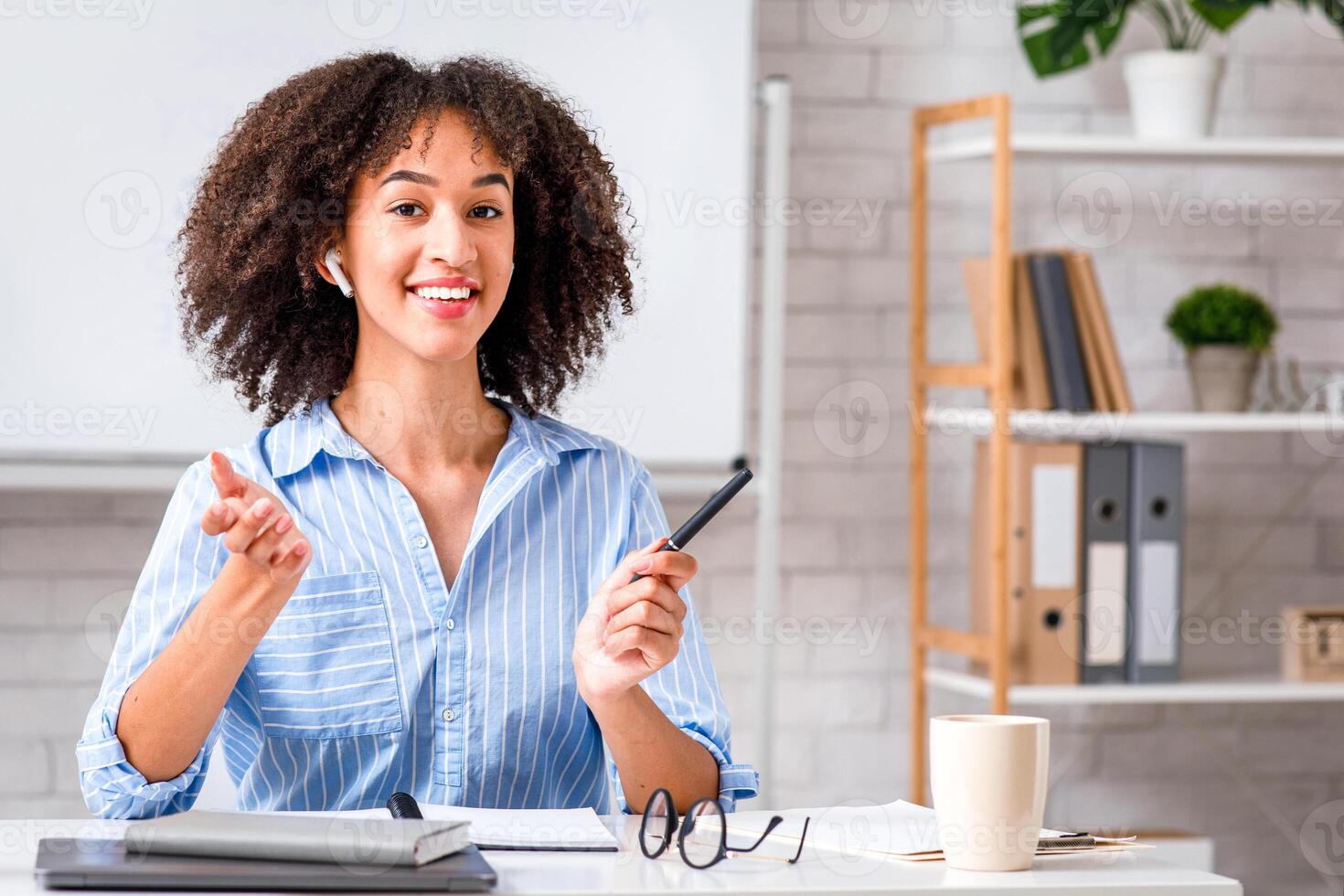 Tutor or teacher of English for students during quarantine and remote lessons. Friendly african american female in casual explains language rules sitting at table with laptop and looking at camera photo