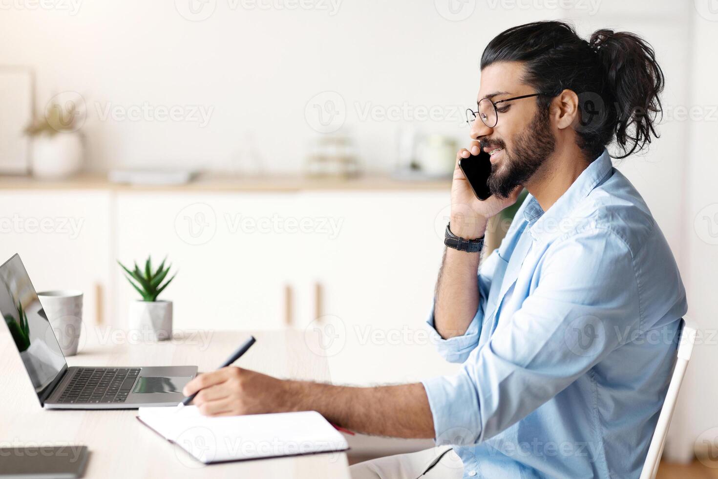 Cheerful Arab Freelancer Guy Talking On Cellphone And Writing Notes To Notepad, Sitting At Desk In Home Office, Speaking On Mobile Phone And Noting Information, Side View With Copy Space photo