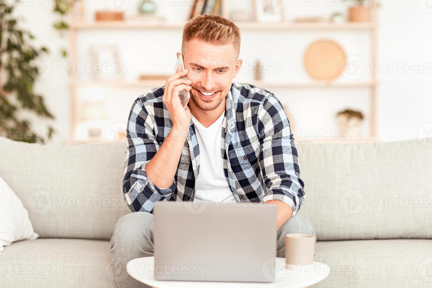 Communication Concept. Smiling millennial man talking on the cellphone using laptop at home office, happy young guy making phone call having mobile conversation looking at computer sitting on sofa photo
