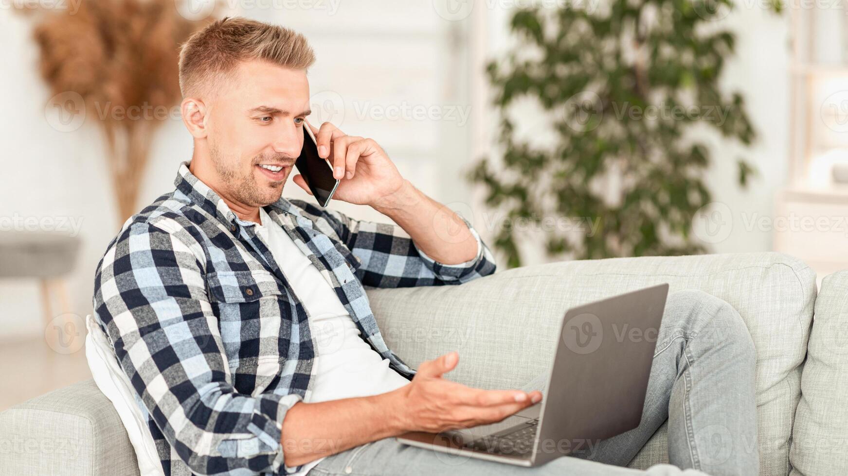 Comfortable Workplace. Young casual guy talking on smart phone and working on laptop, lying on couch photo