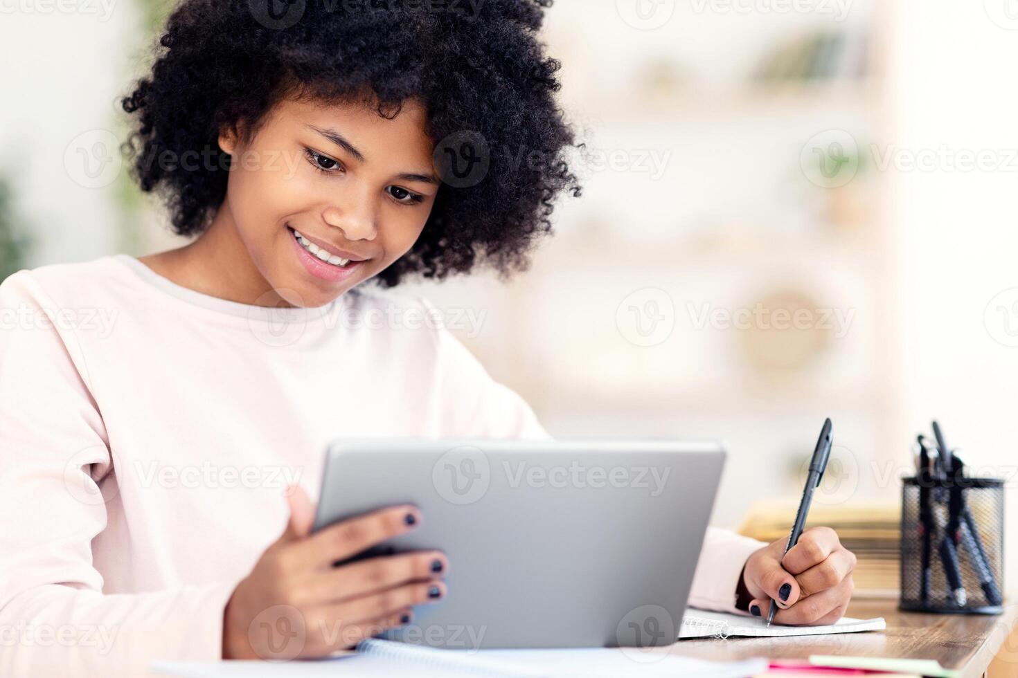 Black Teenager Girl Using Tablet Computer Learning Distantly Online Sitting At Desk At Home. Web Education And E-Learning photo