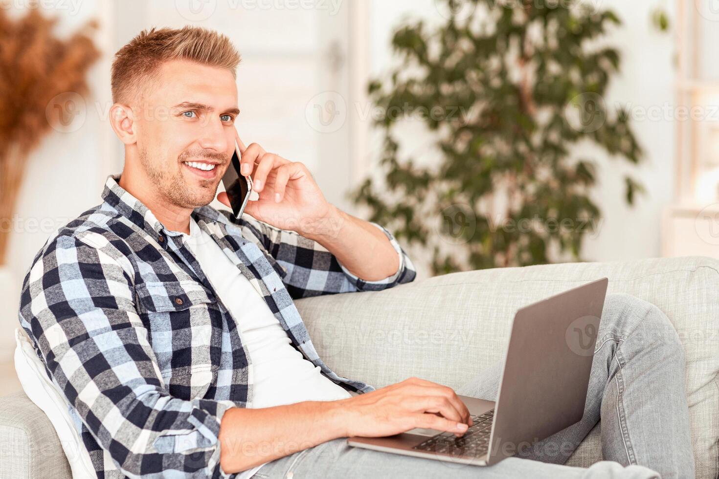 Managing Business Concept. Portrait of confident millennial man talking on cellphone using laptop, resting on the couch in living room. Happy young male making phone call having mobile conversation photo