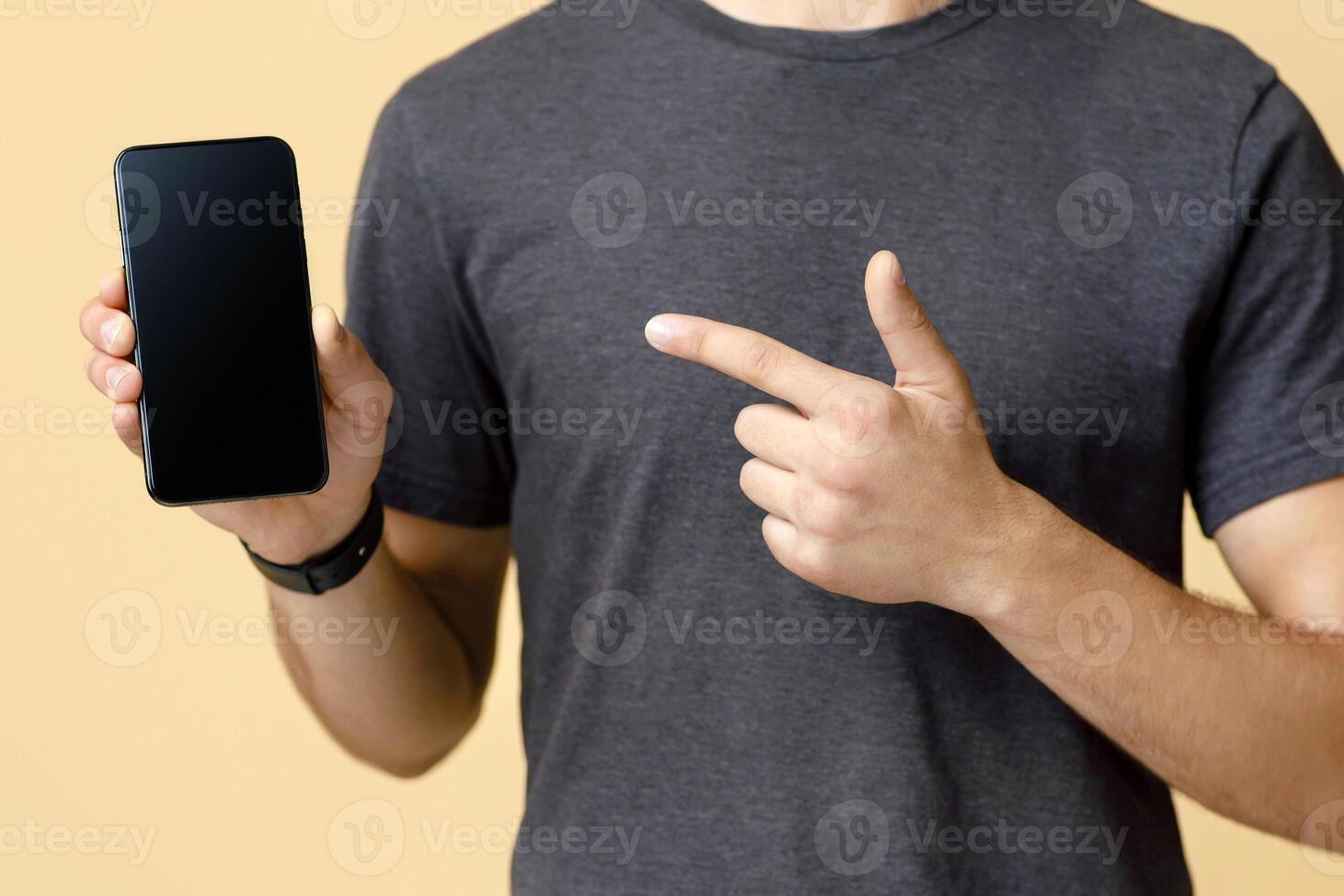 Client advice, gadget review and modern advertising. Man in brown t-shirt, points finger at phone with blank screen, isolated on beige background, free space, cropped, close up, studio shot photo