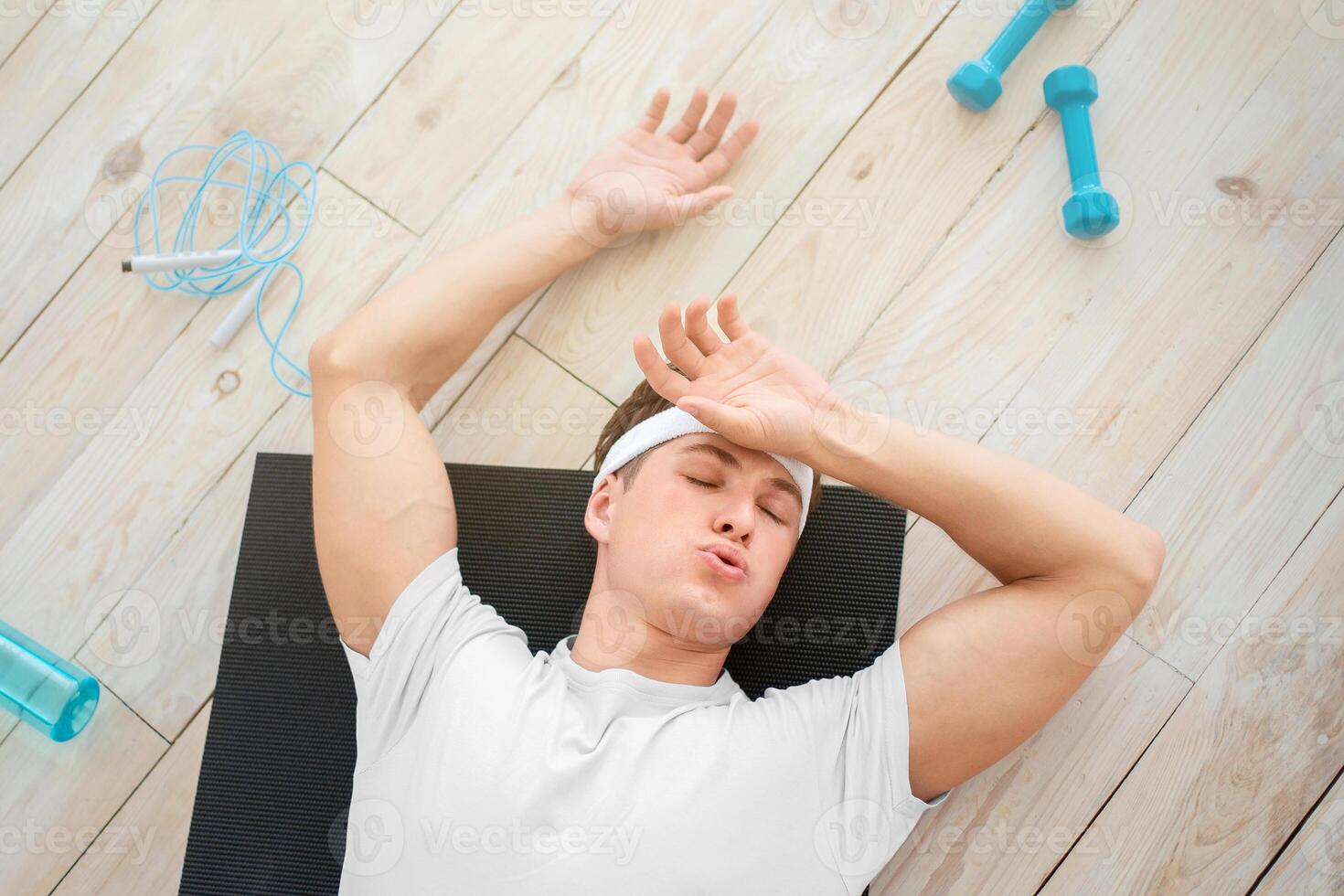 Fatigue after exercising at home. Tired young man wipes sweat, lying on mat on floor with sports tools and empty bottle for water, top view, free space photo
