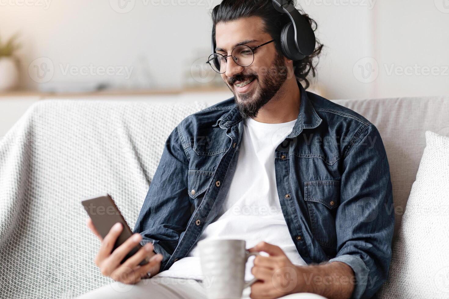 Domestic Pastime. Relaxed Arab Guy Listening Music At Home, Using Smartphone And Wireless Headphones, Drinking Coffee While Resting On Couch In Living Room, Enjoying Weekend Leisure, Free Space photo