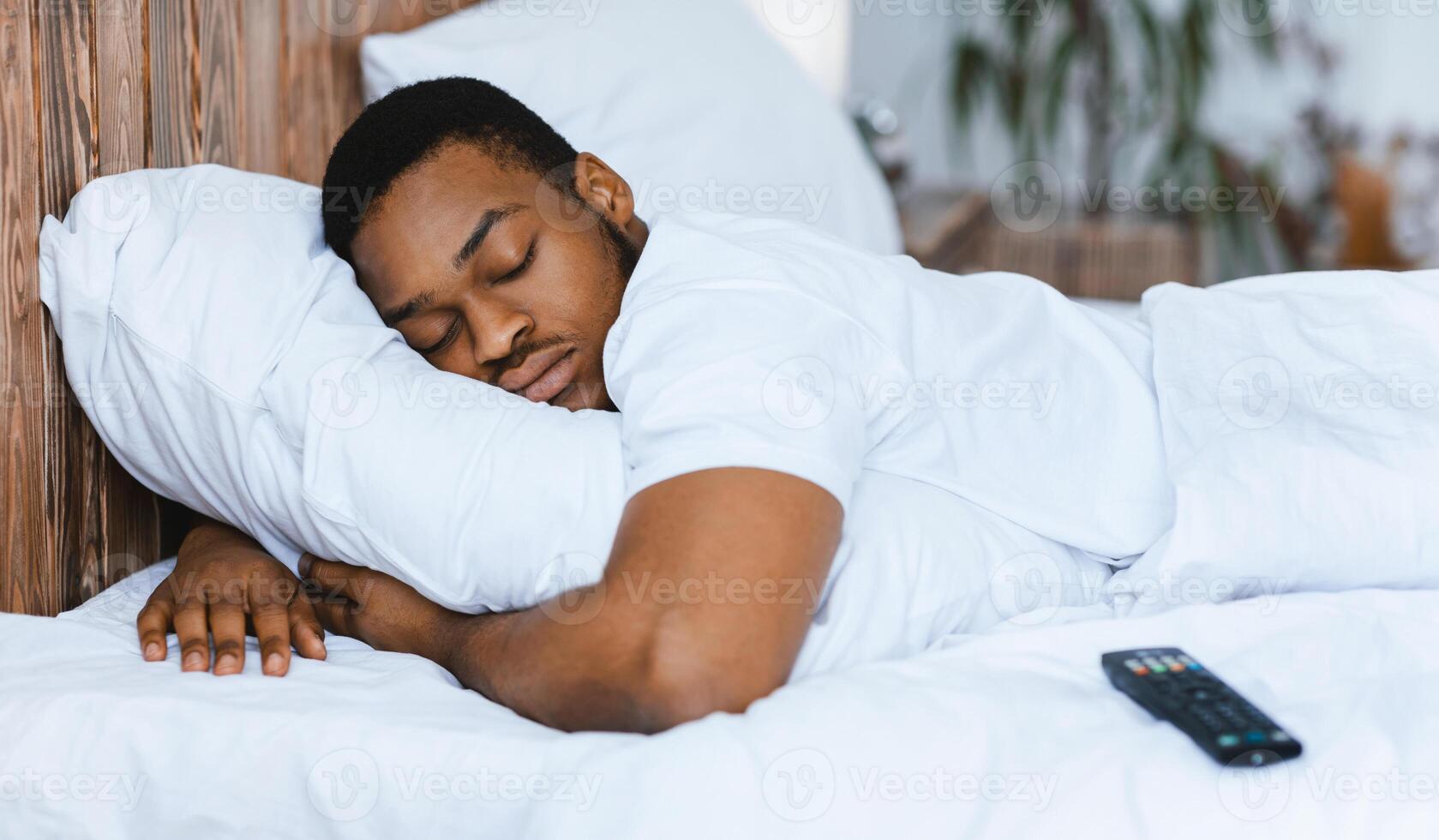 Fast Asleep Black Man Sleeping Embracing Pillow Resting In Bed At Home, Lying Near Remote Control Tired After Bingewatching TV Shows At Night. Rest, Napping And Recreation Concept. photo