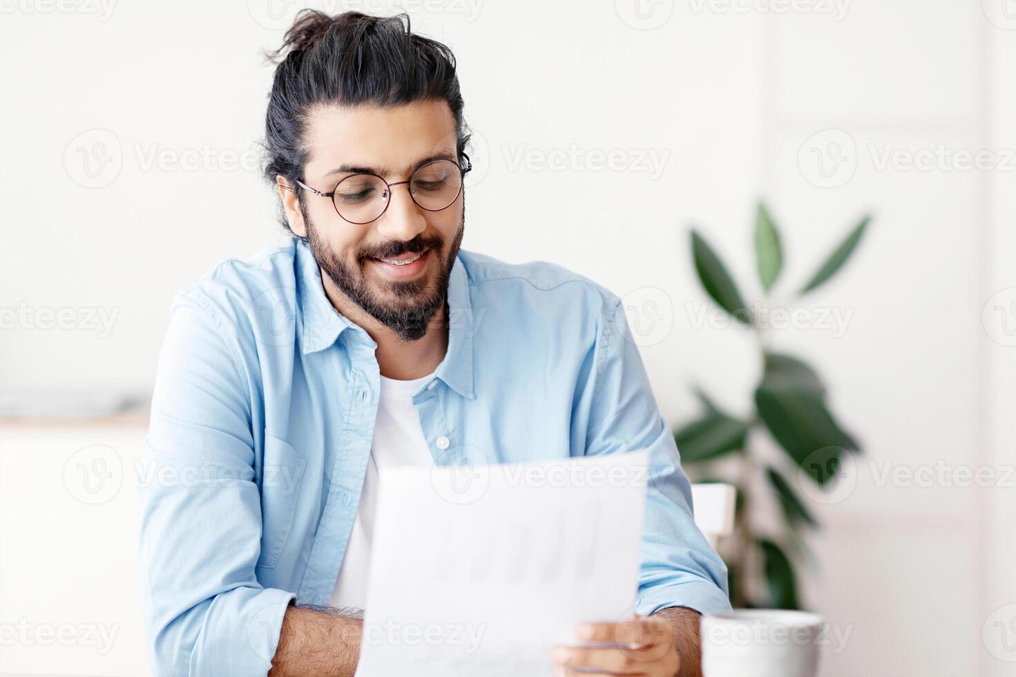 Young Arab Male Self-Entrepreneur Working With Papers At Desk In Home Office, Checking Annual Financial Reports. Eastern Millennial Man Reading Documents, Sitting At Table With Computer, Copy Space photo