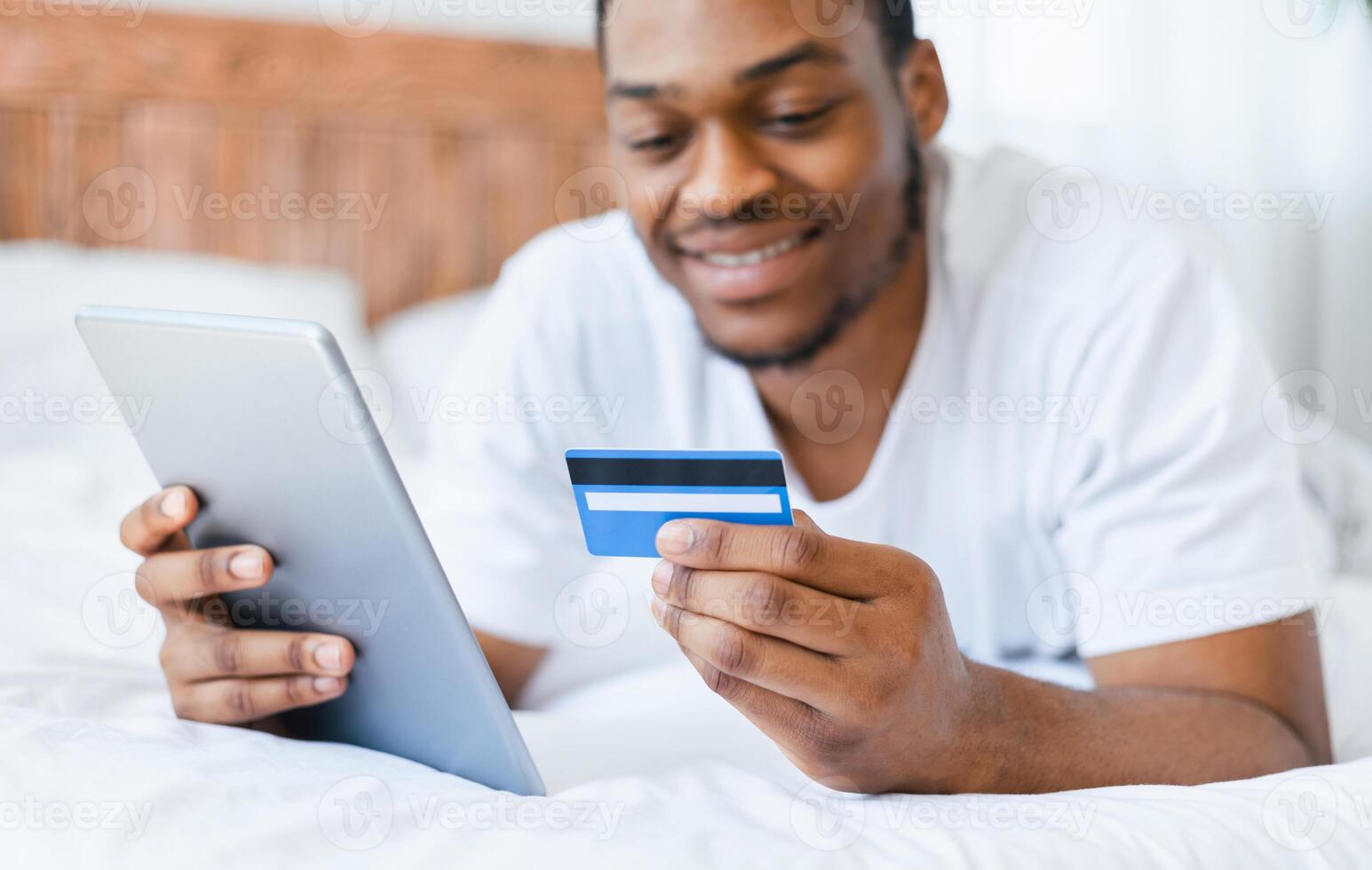 African American Man Shopping Online Using Digital Tablet And Credit Card Lying In Bed At Home. Internet Shop Offers And Sales, E-Commerce Concept. Selective Focus, Shallow Depth photo