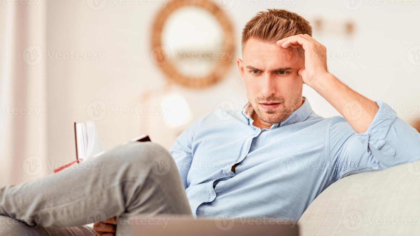 Planning And Project. Portrait of focused pensive man working from home, sitting on couch, holding notebook, looking at laptop. Guy watching webinar, thinking about remote work and his schedule photo