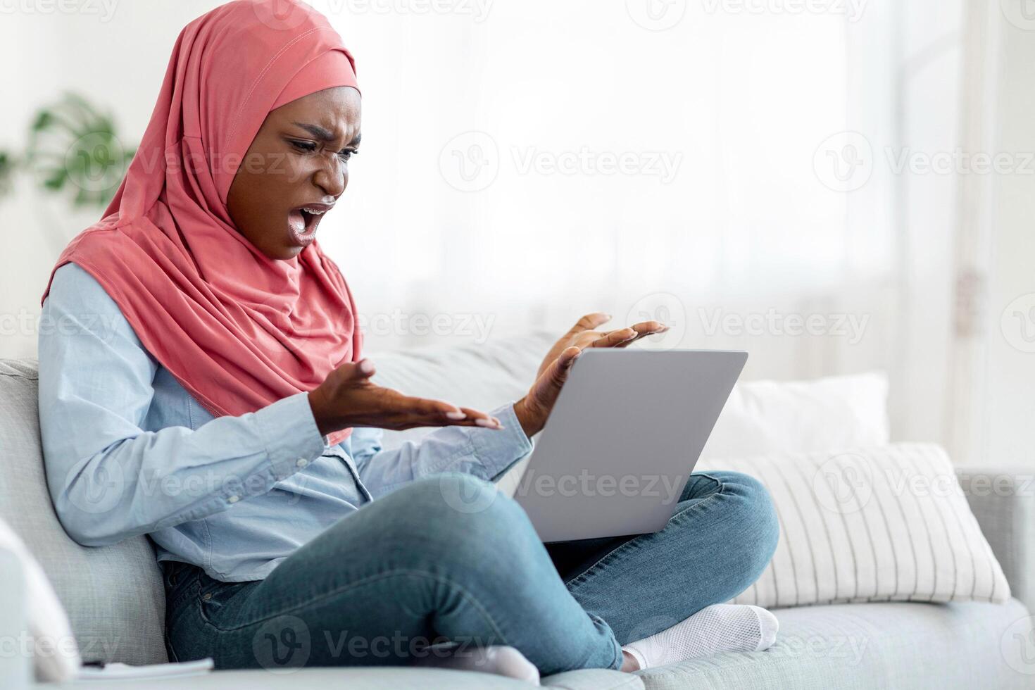Computer Problems. Angry black muslim lady in hijab having troubles with laptop at home, annoyed african woman looking at device screen and emotionally gesturing, having bad internet connection photo