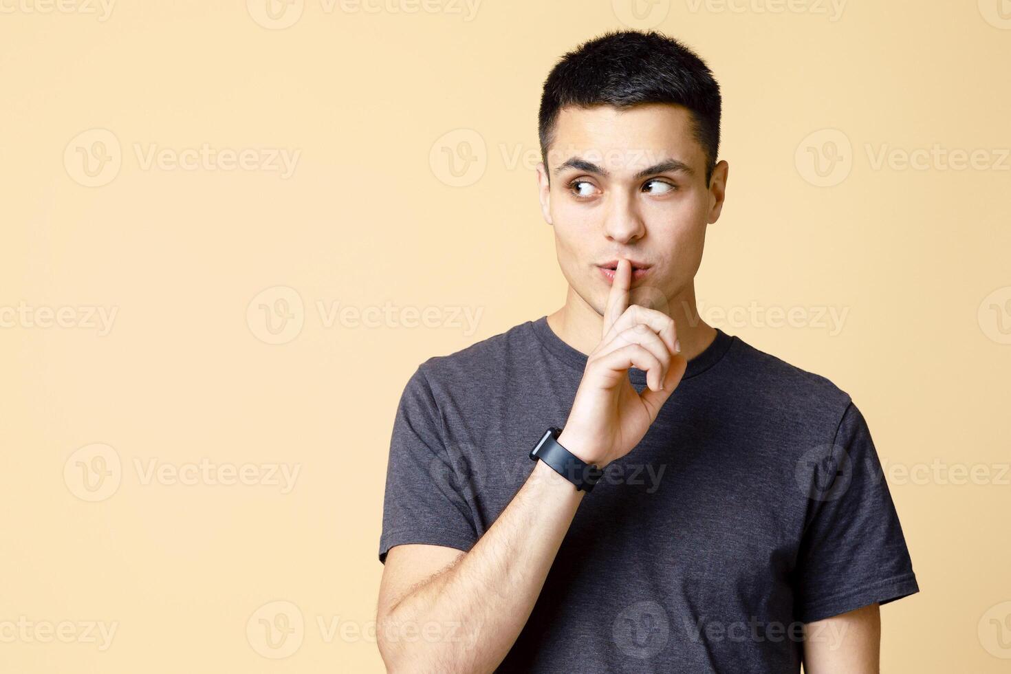 Secret, offer, interesting advertisement and gossip. Mysterious young man student with smart watch shows hand sign silence and looks towards empty space isolated on light sand background, studio shot photo
