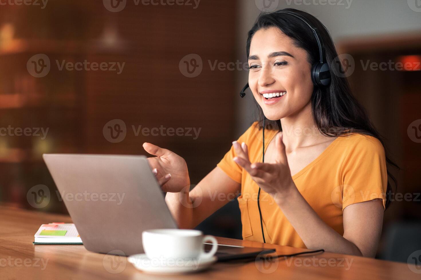 Young arab woman having business conference online, using laptop and headset, cafe interior, free space photo
