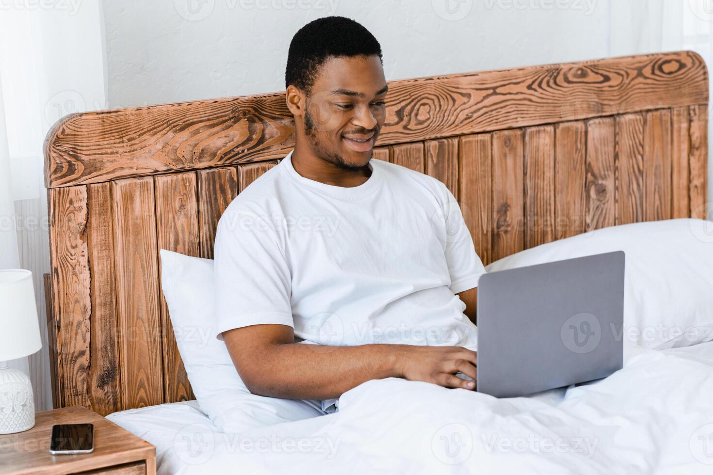 Freelance Career. African American Freelancer Guy Working On Laptop Online Browsing Internet Sitting In Bed At Home. Distant Computer Job Concept. Selective Focus photo