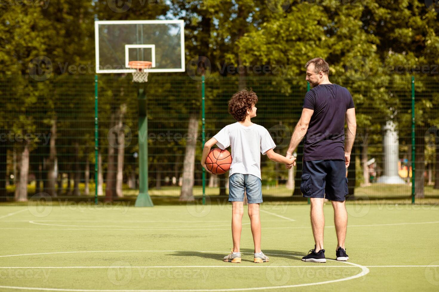 Back view full length portrait of mature man and boy standing on sports field with basket ball, holding hands photo