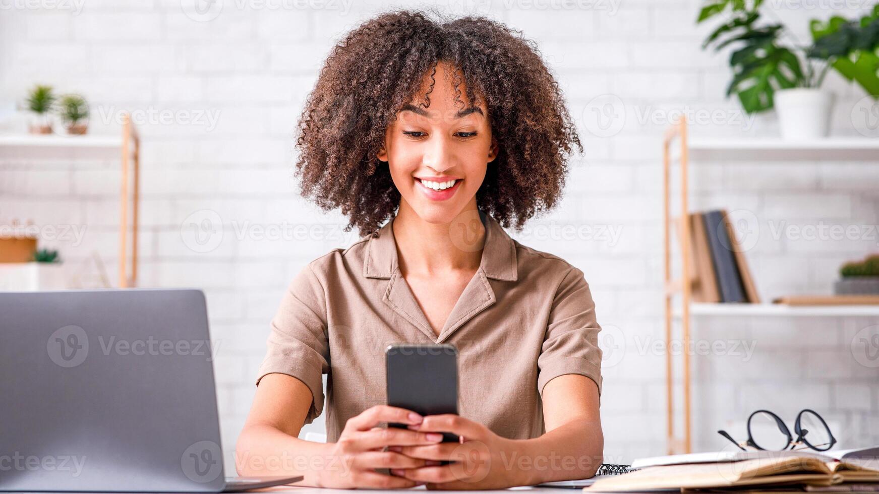 Modern technology for study and work at home during epidemic. Smiling african american woman typing on smartphone, sitting at table with notebook and notepad in living room interior, free space photo