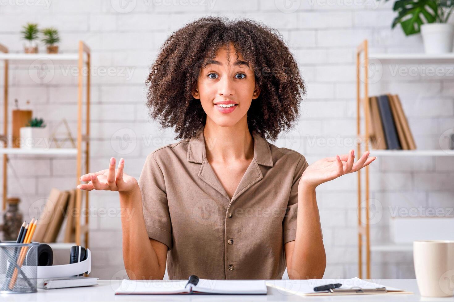 I do not know or maybe. Attractive african american female spreads her arms to the sides and looks at camera, sitting at table with notepad in living room interior and records streaming photo