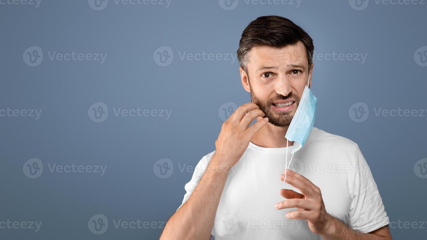 Annoyed bearded man removing protective mask, scratching his irritated face on grey studio background. Man with beard suffering from mask cafe during pandemic, dermatological issues during quarantine photo