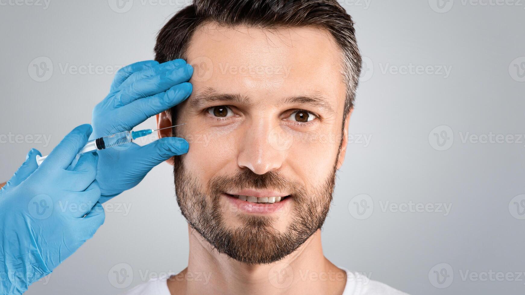 Closeup of middle aged handsome bearded man getting filler for eyes zone at beauty salon or aesthetic clinic, grey studio background. Doctor hands in protective blue gloves injecting filler photo