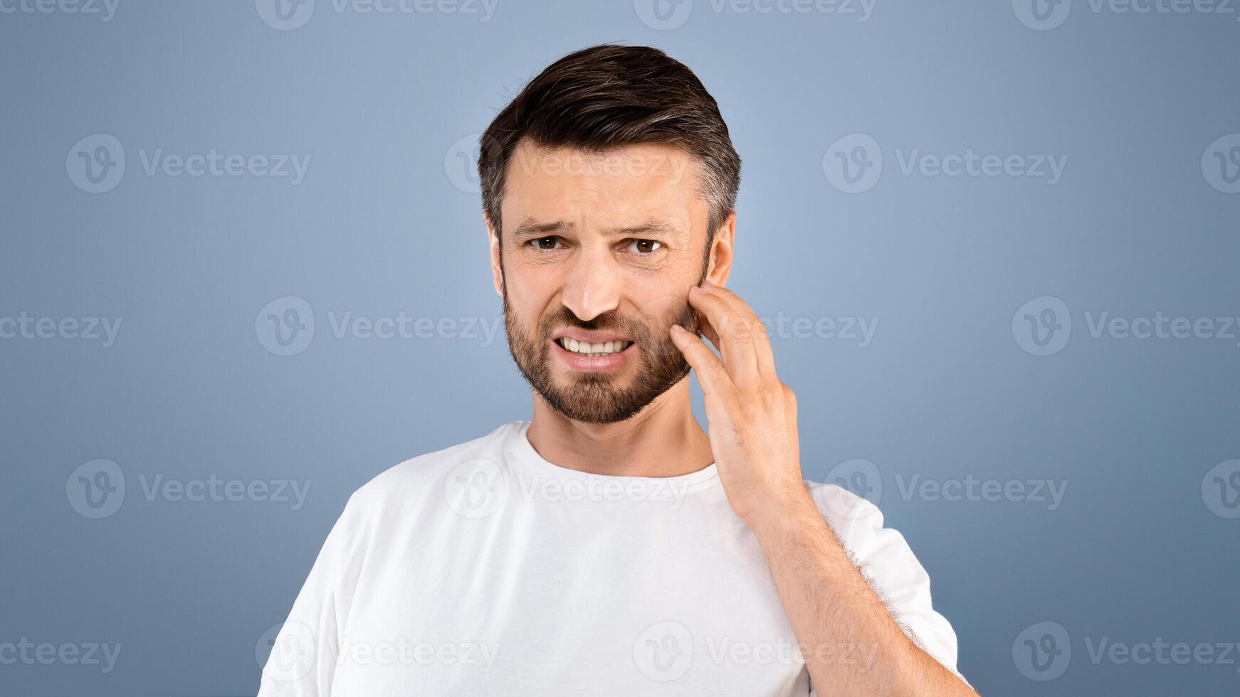 Angry bearded man holding protective mask and scratching irritated face on grey studio background. Man with beard suffering from mask cafe during lockdown, dermatological problems during quarantine photo