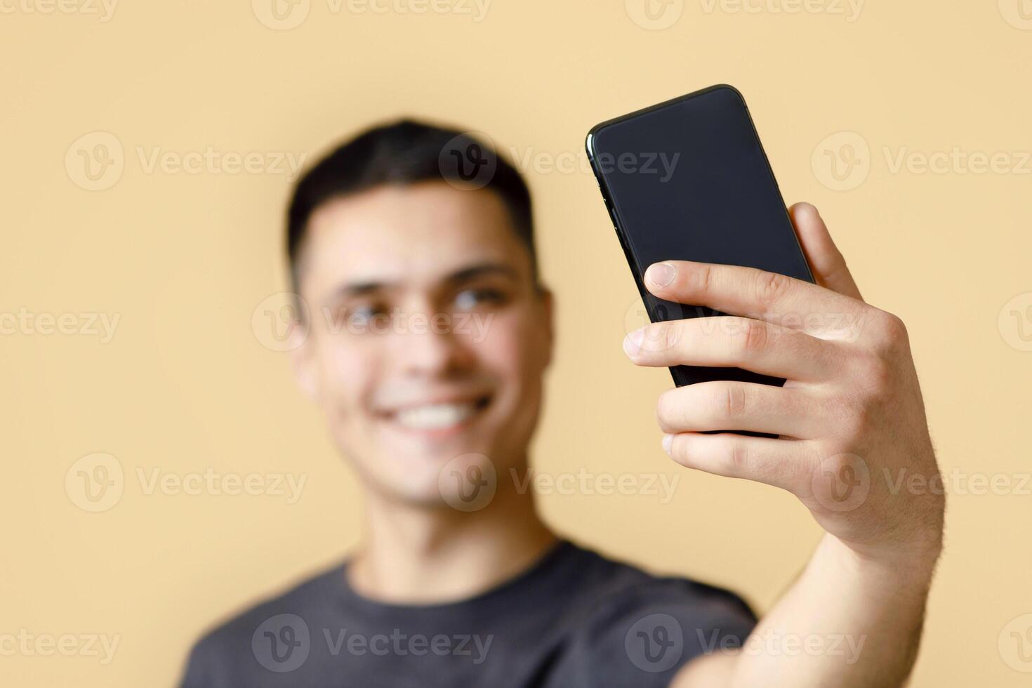 Selfie for blog and social media. Focus on smartphone in hand of smiling young guy blogger student, making photo, isolated on light sand background, blurred, empty space, studio shot photo