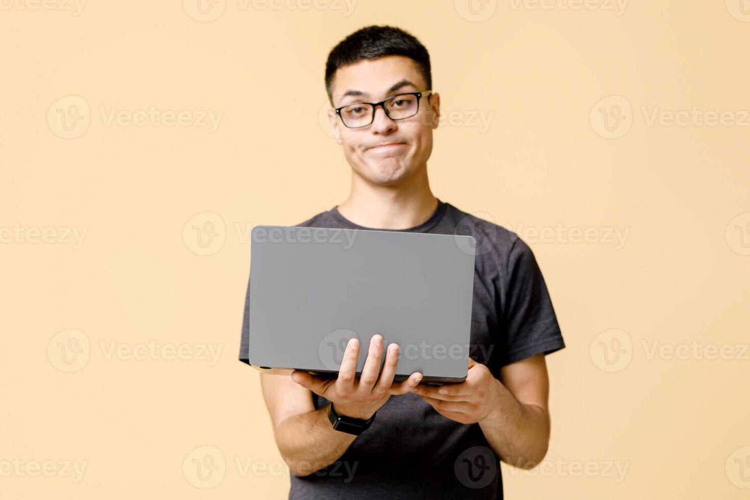 Problems at work, difficulties. Frustrated young guy student with glasses looking at camera and holds laptop, isolated on beige background, studio shot, free space photo