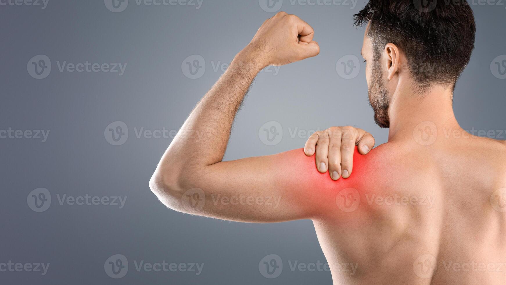 Back view of topless man suffering from shoulder pain injury, rubbing sore zone over grey studio background. Healthcare and medical concept, sports traumatology and orthopedics photo