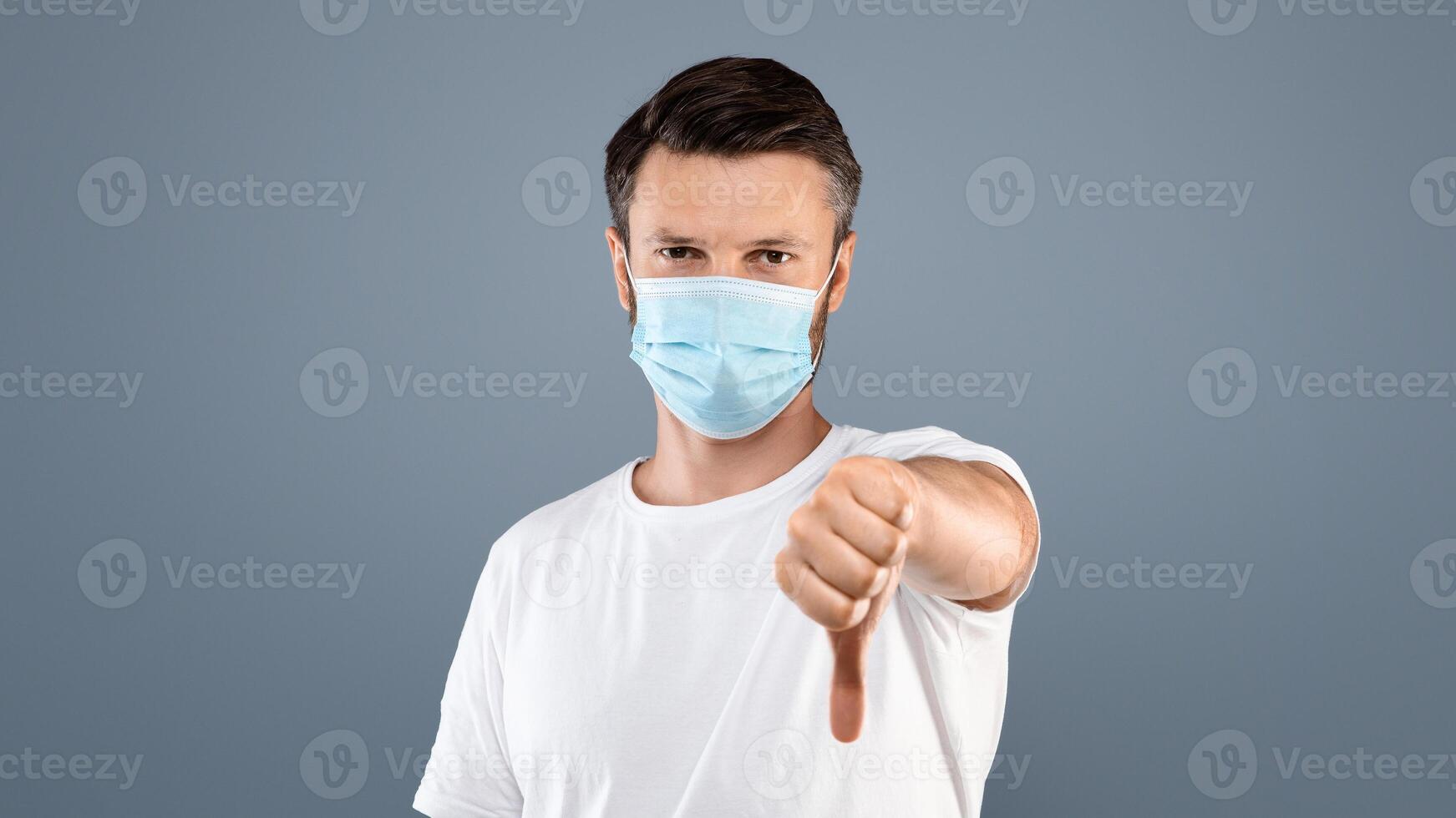 Young man in medical protective mask showing thumb down over gray studio background. Man in white t-shirt with blue medical mask showing dislike gesture, stay safe and healthy during pandemic photo