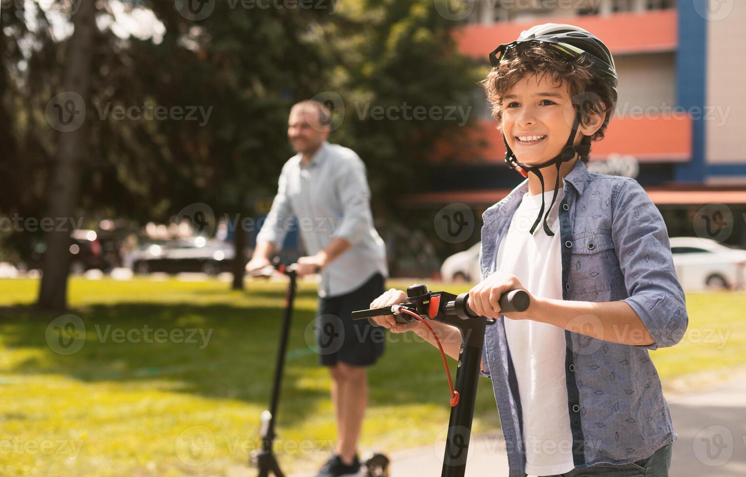 Portrait of happy curly boy in protective helmet having ride on motorized scooter with father in blurred background photo
