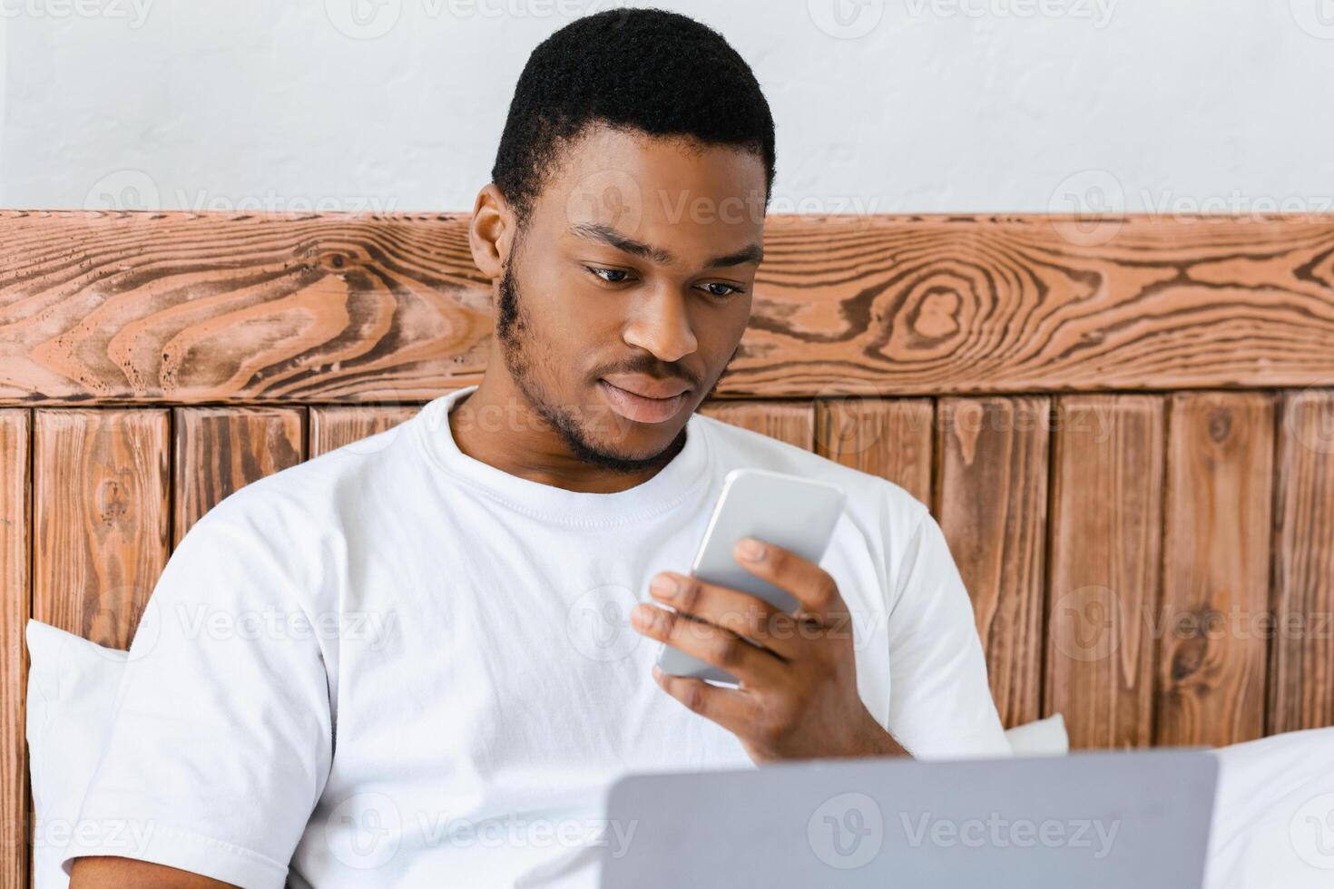 African American Freelancer Man Texting On Phone Using Laptop Computer Working Sitting In Bed At Home. Always In Touch, Freelance Lifestyle And Distant Work Concept. Selective Focus photo