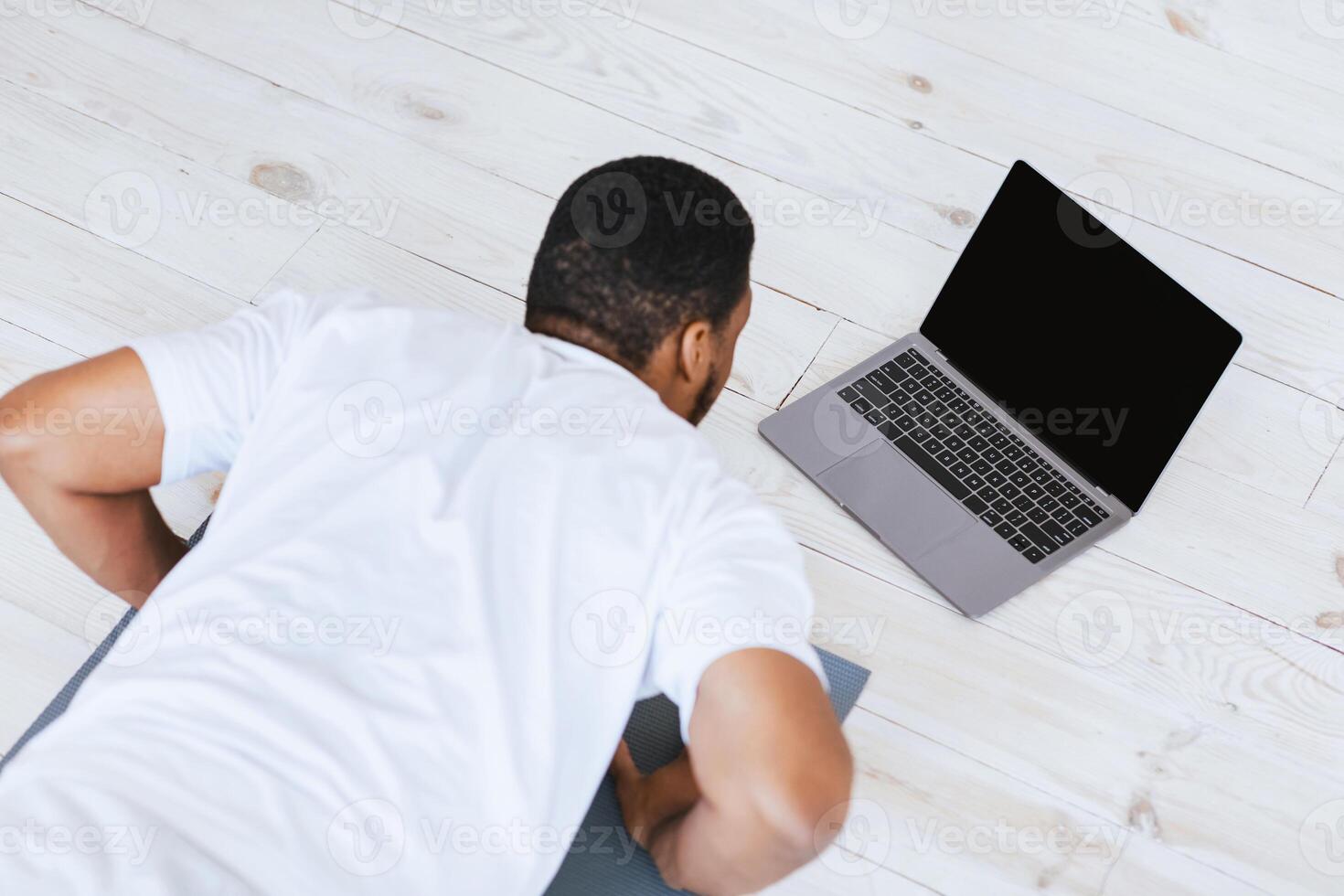 Online Training. African Man At Laptop With Empty Screen Exercising Doing Plank Exercise During Workout At Home. Internet Fitness Classes Advertisement. Mockup, High-Angle Shot, Selective Focus photo