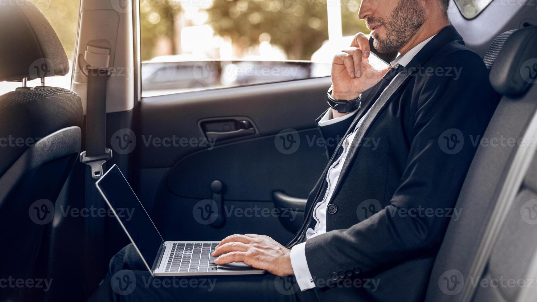 Busy entrepreneur working on laptop, talking on phone while going to airport by car, side view photo