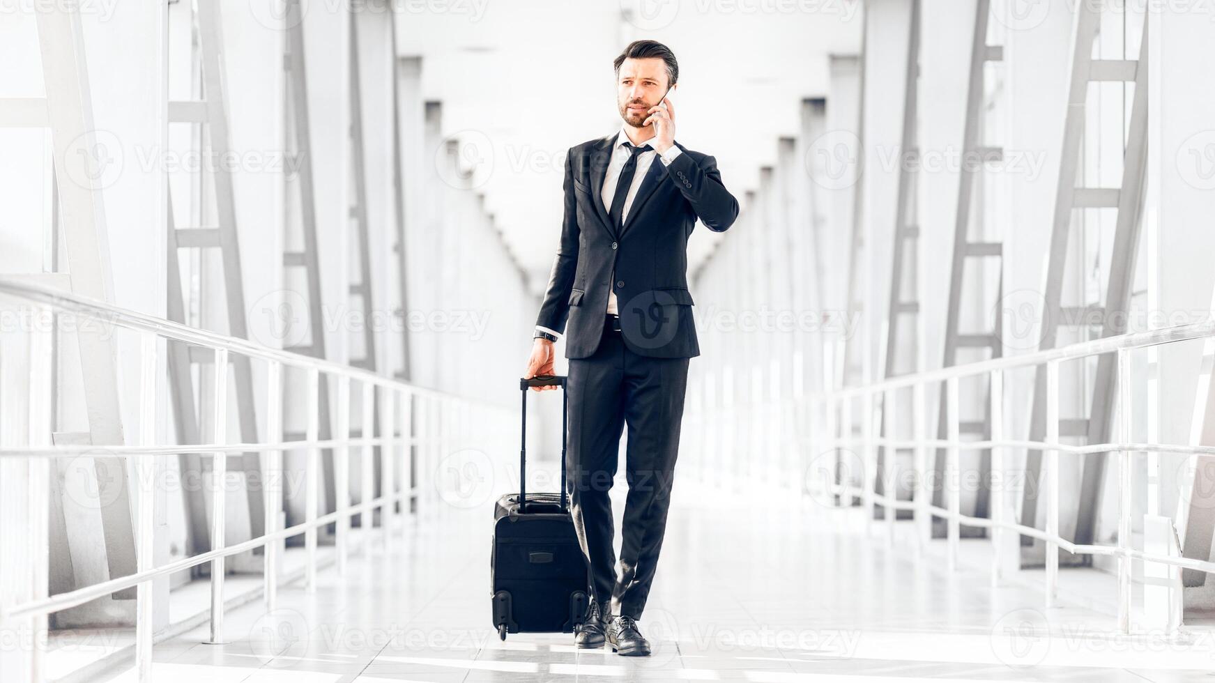 Young businessman talking on phone, walking with his luggage by airport, empty space photo