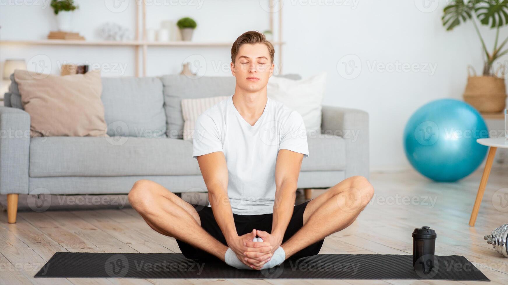 Modern sports at home in self isolation during quarantine. Guy with closed eyes meditates or practices yoga in interior of living room, free space photo