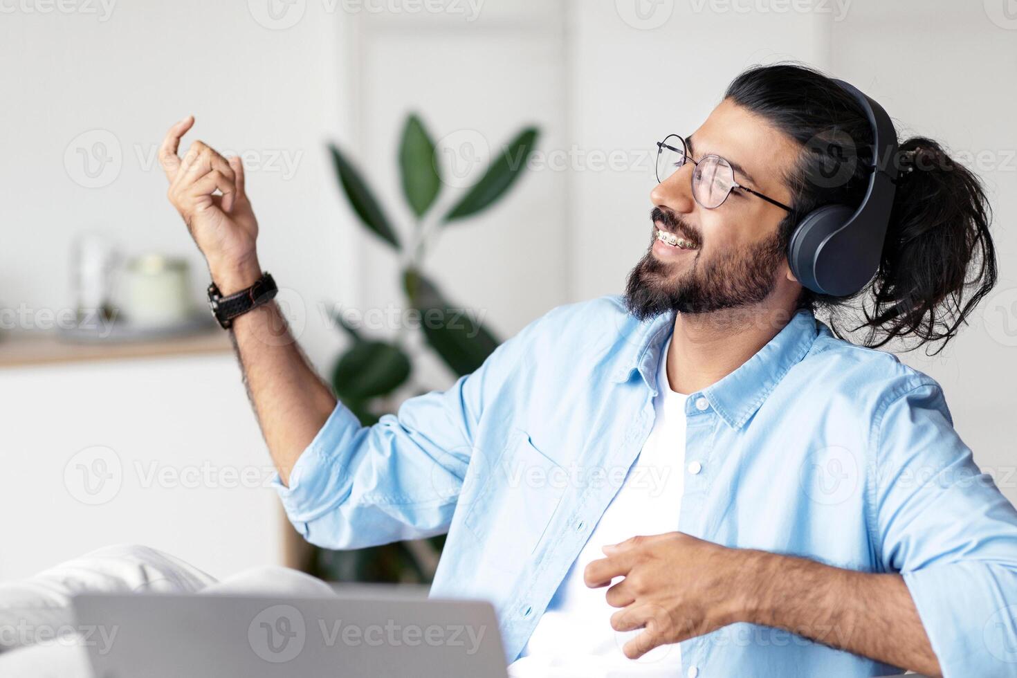 Be Yourself. Joyful weatern freelancer guy listening music in headphones and playing virtual guitar, relaxing at desk in home office, young indian man having fun and enjoying working remotely photo