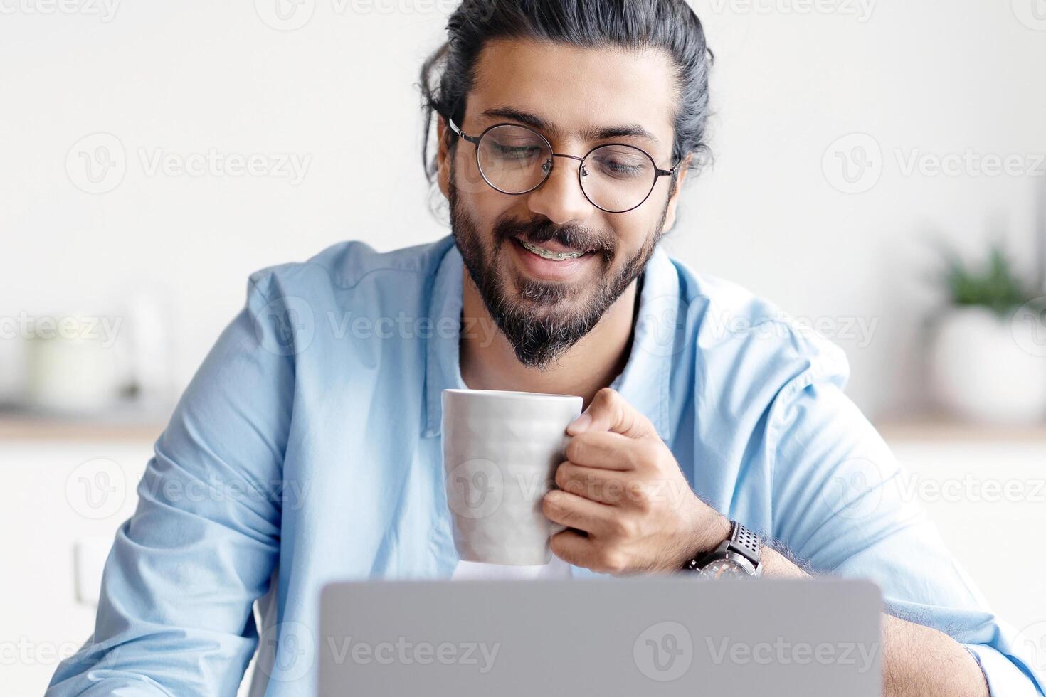 Young Smiling Arab Freelancer Guy Using Laptop And Drinking Coffee At Home, Relaxing With Hot Drink While Working Remotely Online On Computer, Enjoying Distance Job Opportunities, Free Space photo