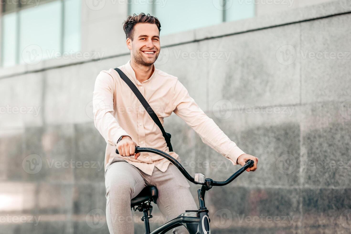 A young man rides his bike down a city street, wearing casual clothes and a smile. The urban backdrop features smooth gray walls and modern architecture, reflecting a vibrant lifestyle. photo