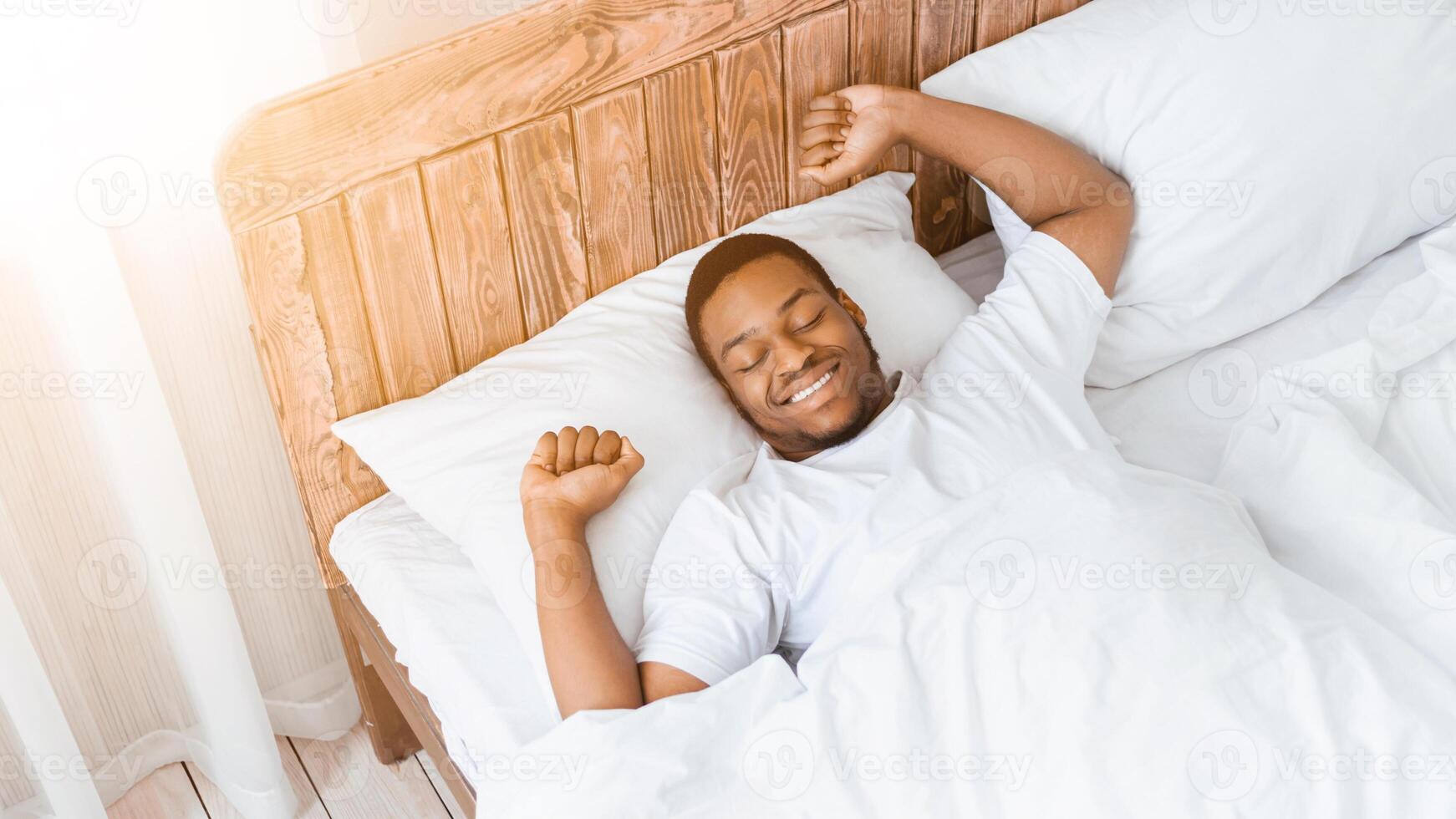 Wellslept African American Man Waking Up Stretching Hands Lying In Bed Smiling With Eyes Closed At Home. Awakening After Good Sleep In The Morning, Rest And Recreation Concept. High-Angle, Panorama photo