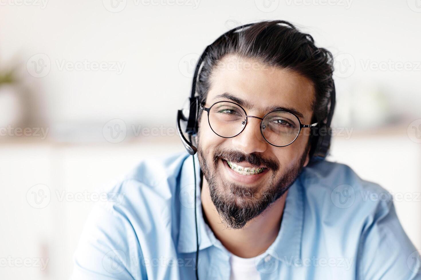 Call Center Operator. Closeup portrait of positive indian man customer service representative in headset, handsome western guy hotline manager looking at camera and smiling, selective focus photo