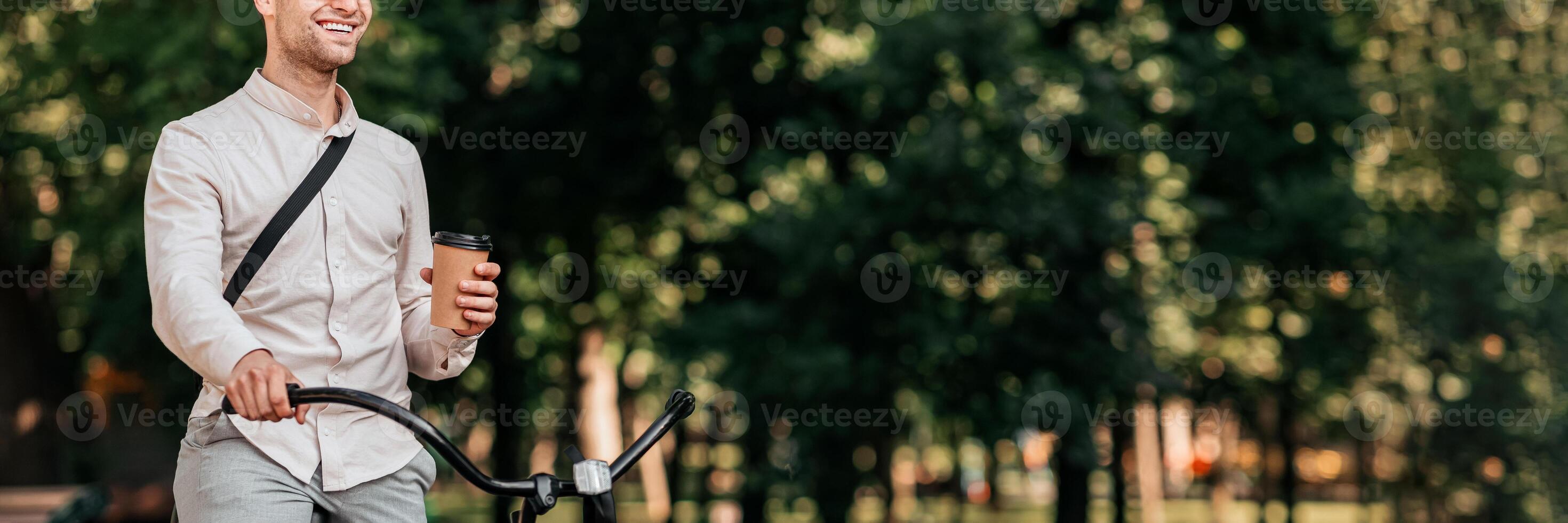 A young man smiles as he rides his bike down an urban path, holding a coffee cup in his hand. This lifestyle moment captures the joy of city life and outdoor activity, web-banner photo