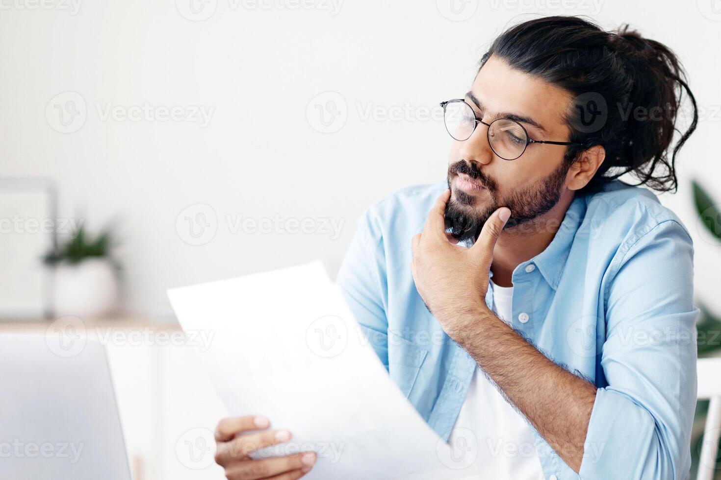Young Arab Male Entrepreneur Working With Papers At Desk In Office, Checking Financial Reports, Millennial Eastern Guy Reading Documents And Touching Chin With Pensive Face Expression, Free Space photo