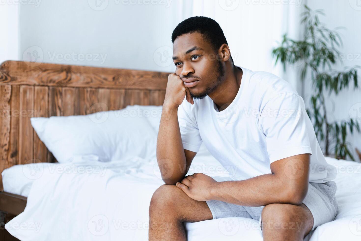 Unhappy Black Guy Sitting On Bed In Bedroom At Home. Depressed African American Man Thinking Of Life Problems. Stress, Apathy And Male Depression Concept. Free Space For Text photo