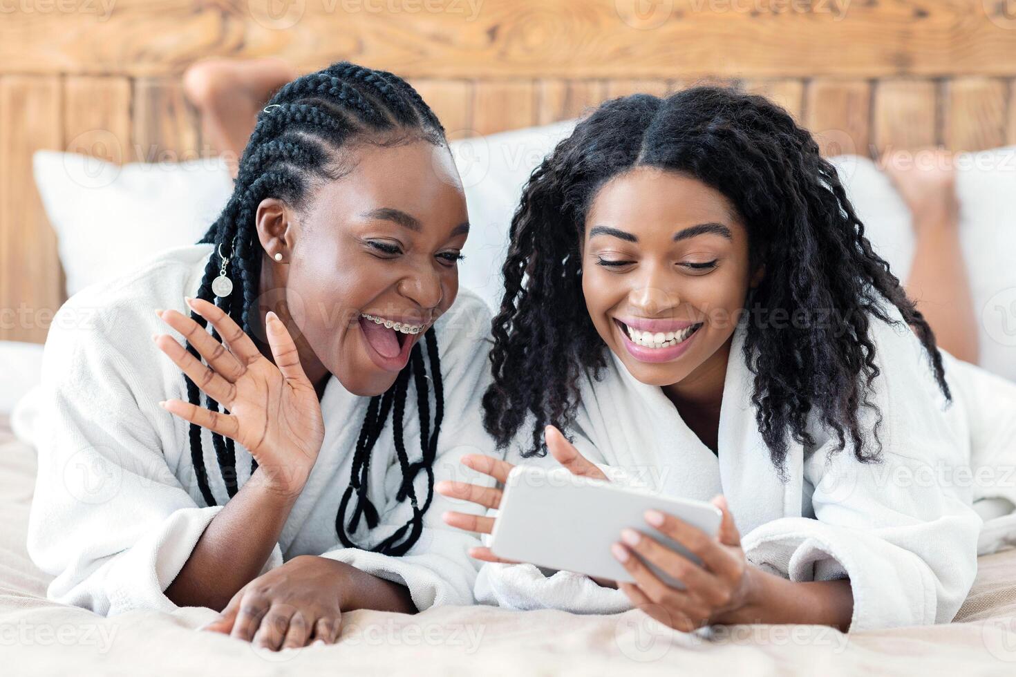 Happy african american young women in bathrobes laying on bed and calling their girlfriend, using mobile phone. Cheerful black girlfriends looking at smartphone screen and waving, having videocall photo