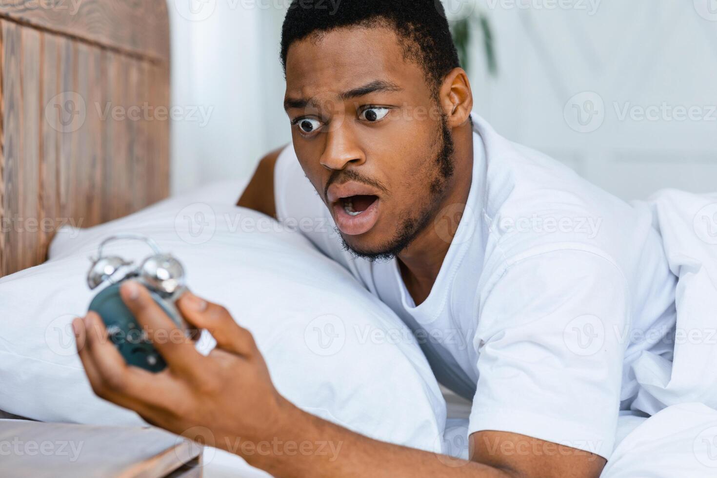 Oversleeping Concept. Shocked Black Guy Looking At Broken Alarm Clock Waking Up Late For Work In The Morning, Lying In Bed At Home. Awakening Problem and Tardiness, Bad Morning photo