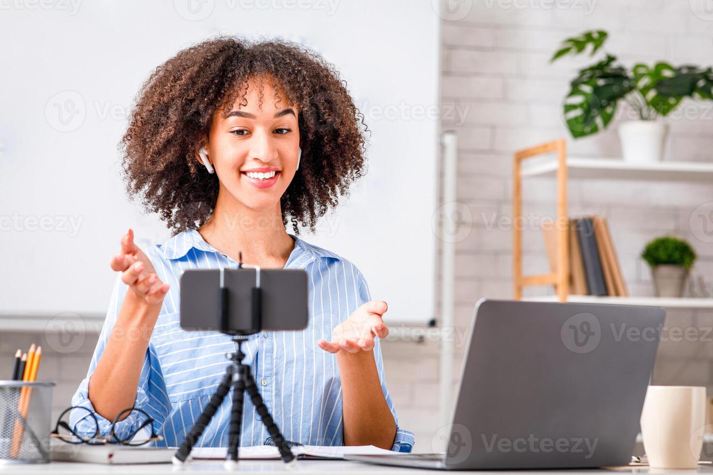 Recording of English lesson with tutor during covid-19 epidemic. Smiling african american lady with wireless headphones makes on web camera, at table with laptop in living room photo