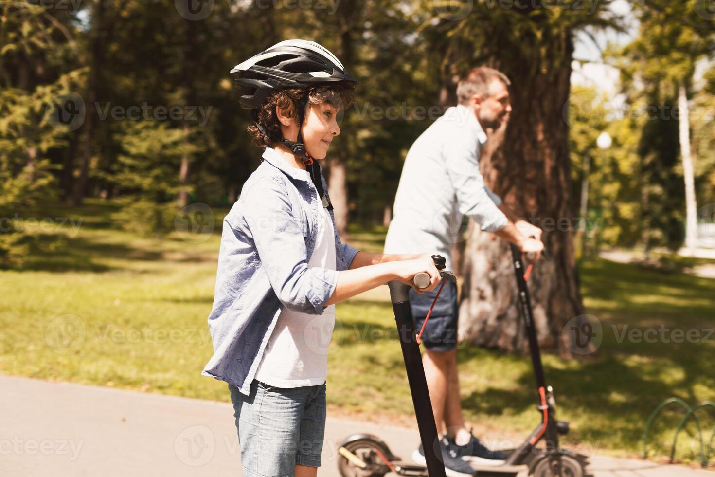 Sharing Service. Cute curly boy riding electric scooters with his daddy in nature, side view, selective focus photo
