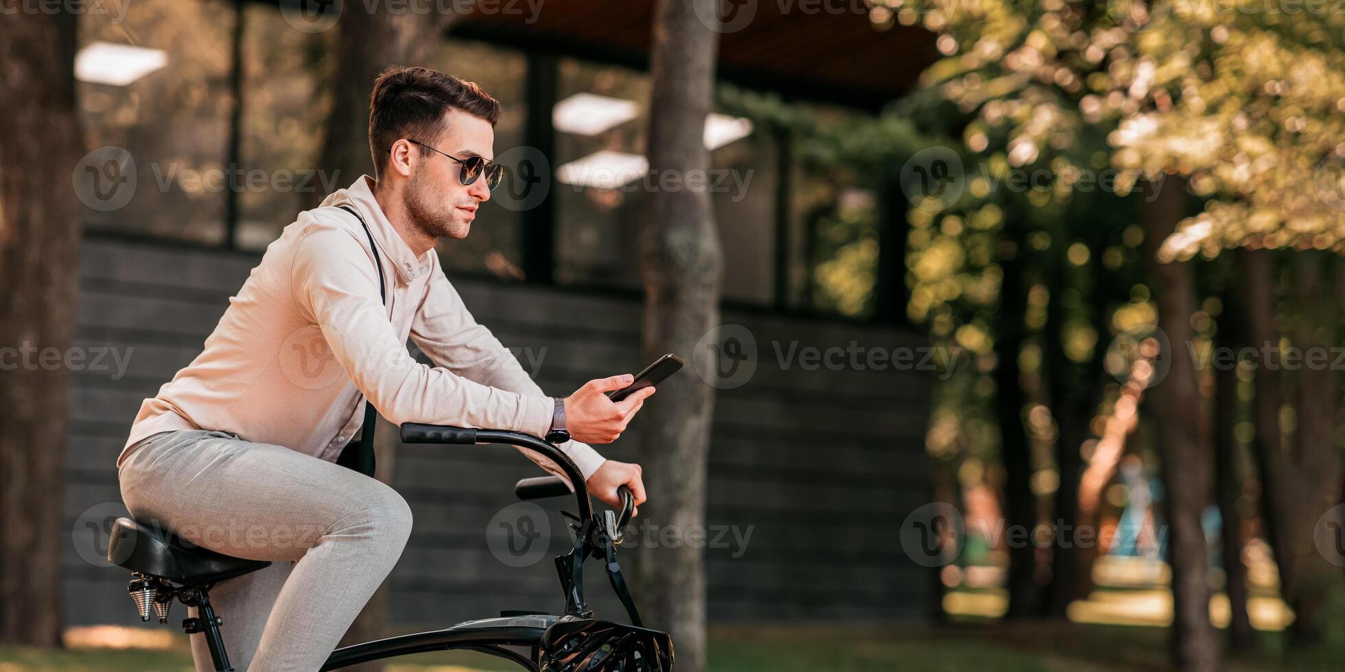 Tries to find his best road during stop with gps online map. Concentrated handsome millennial man in sunglasses looks at smartphone, sits on modern bicycle in park, outdoor, side view, free space photo